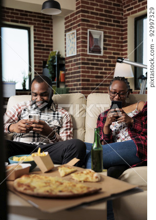Cheerful couple browsing internet on mobile phone and watching tv, eating takeaway fast food from takeout delivery. Dinner meal package to eat and watch movie on television together. Cheerful couple browsing internet on mobile phone and watching tv, eating takeaway fast food from takeout delivery. Dinner meal package to eat and watch movie on television together. 92997329