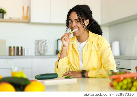 Happy black woman cooking fresh vegetable salad and biting cucumber while preparing lunch in kitchen Happy black woman cooking fresh vegetable salad and biting cucumber while preparing lunch in kitchen 92997330