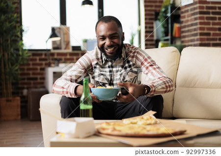 Smiling person laughing at comedy movie on television and eating chips from bowl, having takeout delivery meal and beer on table. Having fun with film on tv and enjoying takeaway fast food. 92997336