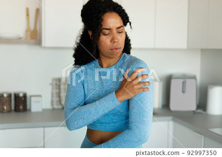 Sporty black woman putting her hand on injury shoulder, having pain, standing in kitchen interior at home 92997350