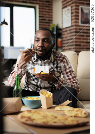 Relaxed man enjoying noodles package from takeaway delivery, eating food with chopsticks in front of television movie. Watching film on tv channel program, having fun with takeout meal. 92997369