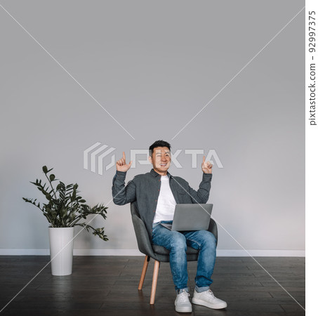 Glad smiling middle aged chinese male with laptop making victory gesture with hands sit in armchair 92997375