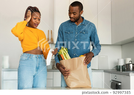 Shocked black spouses with paper bag with food checking grocery bill, woman touching her head, standing in kitchen Shocked black spouses with paper bag with food checking grocery bill, woman touching her head, standing in kitchen 92997385