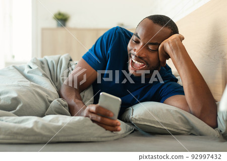 Cheerful Black Man Using Smartphone Lying In Bed In Bedroom 92997432
