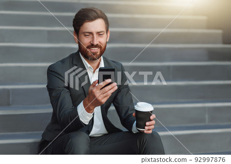 Smiling office worker in suit sitting on stairs with coffee during break and using phone 92997876
