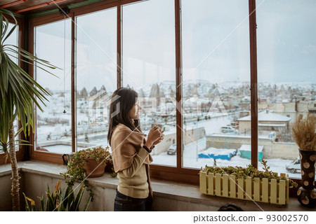 woman enjoy coffee standing next to a window with beautiful view of snowy landscape woman enjoy coffee standing next to a window with beautiful view of snowy landscape 93002570