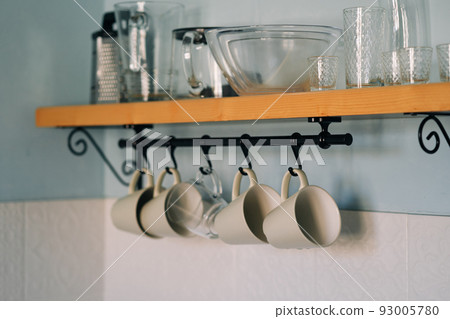 Kitchen open wooden shelf with dishes, glasses and mugs. Mugs are hanging from hooks. Cozy interior in a country house. Kitchen open wooden shelf with dishes, glasses and mugs. Mugs are hanging from hooks. Cozy interior in a country house. 93005780