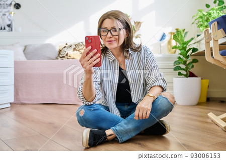 Middle aged woman sitting on floor at home with smartphone Middle aged woman sitting on floor at home with smartphone 93006153