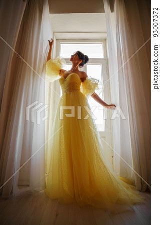 A woman's silhouette in a golden luxurious dress against the background of a window holds a curtain with her hands. Elegant lady in a yellow long silk dress with bare back, back view. A woman's silhouette in a golden luxurious dress against the background of a window holds a curtain with her hands. Elegant lady in a yellow long silk dress with bare back, back view. 93007372