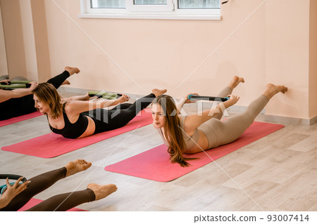 A group of six athletic women doing pilates or yoga on pink mats in front of a window in a beige loft studio interior. Teamwork, good mood and healthy lifestyle concept. A group of six athletic women doing pilates or yoga on pink mats in front of a window in a beige loft studio interior. Teamwork, good mood and healthy lifestyle concept. 93007414