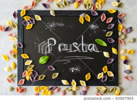 Multi-coloured Italian pasta conchiglie or seashells with basil leaves on chalk board. Word Pasta wrote with chalk Multi-coloured Italian pasta conchiglie or seashells with basil leaves on chalk board. Word Pasta wrote with chalk 93007905