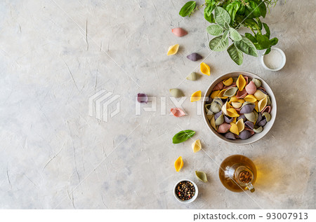 Multi-coloured Italian pasta conchiglie or seashells in bowl, olive oil, spices, herbs. Flatly with copy space.  93007913