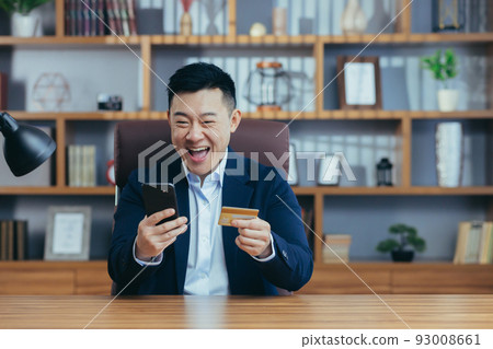 Young handsome asian man in suit in the office at work holding a credit card and shopping, ordering online from the phone, happy Young handsome asian man in suit in the office at work holding a credit card and shopping, ordering online from the phone, happy 93008661