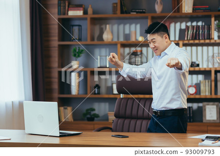 Happy businessman dancing and celebrating victory sitting at a table in a classic office, Asian working at computer Happy businessman dancing and celebrating victory sitting at a table in a classic office, Asian working at computer 93009793