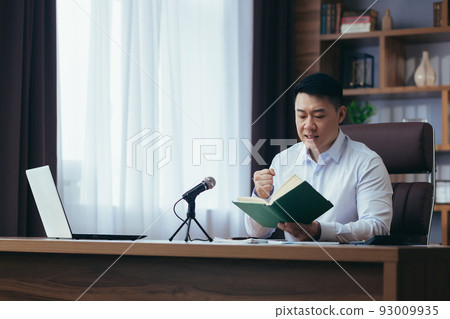 Asian preacher reads the bible online, records a podcast, sits at a table and looks at the camera, uses a microphone, holds his hand in his heart, online radio broadcast Asian preacher reads the bible online, records a podcast, sits at a table and looks at the camera, uses a microphone, holds his hand in his heart, online radio broadcast 93009935
