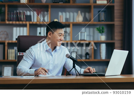 Focused smiling young businessman using professional microphone recording audio podcast, voice acting or streaming training webinar, Asian looking at laptop screen. 93010159