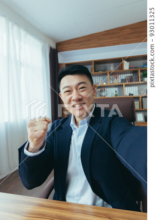 Cheerful and funny Asian businessman looks at the camera of a smartphone, talks on a video call, a man works in the office, waves a congratulatory gesture 93011325