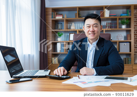 Portrait of young handsome asian man, businessman, accountant. Works with a laptop, calculator, documents, in a suit at the table in the office. Smiles 93011629