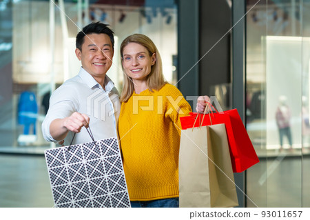 Young asian man and woman couple in supermarket shopping, looking at camera and smiling, holding colored packages with goods 93011657
