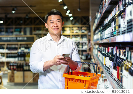 Asian man in supermarket, uses phone and app to buy and choose wine in store, looks at camera and smiles Asian man in supermarket, uses phone and app to buy and choose wine in store, looks at camera and smiles 93013051