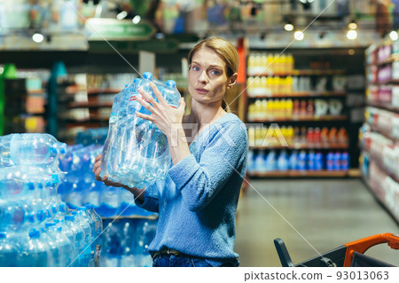Portrait of scared woman shopper in supermarket buying water in plastic bottles and looking scared at camera Portrait of scared woman shopper in supermarket buying water in plastic bottles and looking scared at camera 93013063