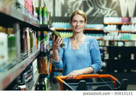 Portrait of a happy woman shopper in the department of household chemicals, woman holding and unwrapping shampoo for hair care, a buyer in a large supermarket Portrait of a happy woman shopper in the department of household chemicals, woman holding and unwrapping shampoo for hair care, a buyer in a large supermarket 93013131