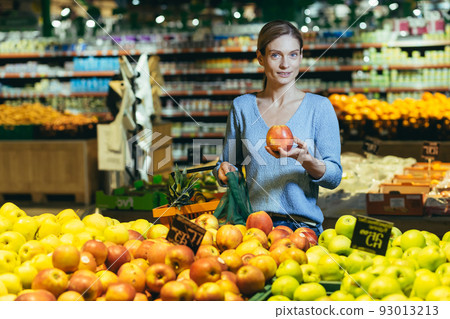 woman picks chooses fruits vegetables on the counter in supermarket. Female housewife shopping in market standing near vegetable department store with a basket in hands. Examines apple 93013213