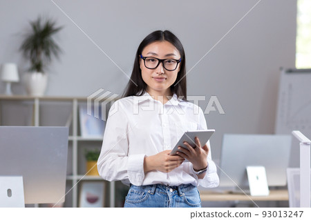 Portrait of young beautiful Asian female student in glasses, white shirt and jeans. Standing in the office, holding a tablet in his hands, studying, looking at the camera, smiling 93013247