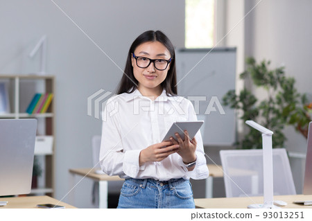 Portrait of young beautiful Asian female student in glasses, white shirt and jeans. Standing in the office, holding a tablet in his hands, studying, looking at the camera, smiling Portrait of young beautiful Asian female student in glasses, white shirt and jeans. Standing in the office, holding a tablet in his hands, studying, looking at the camera, smiling 93013251