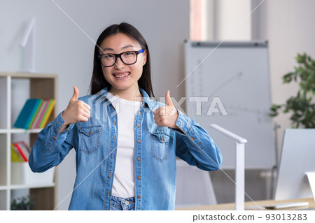 Portrait of young asian student girl in glasses and denim clothes, standing, showing super with two hands, showing success with fingers, looking at camera, smiling. Portrait of young asian student girl in glasses and denim clothes, standing, showing super with two hands, showing success with fingers, looking at camera, smiling. 93013283