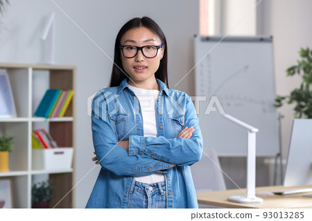 Portrait of a young Asian student girl in glasses and denim clothes, standing, arms crossed, looking at the camera, smiling 93013285