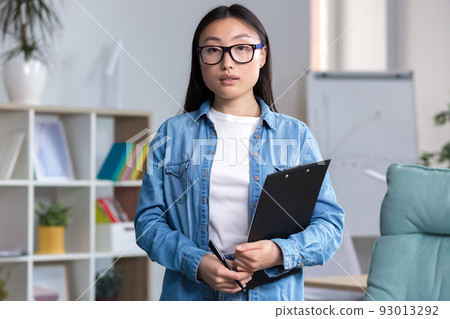 Young Asian woman in glasses, intern, assistant, secretary for the first time at a workplace in the office. He holds a folder in his hands, looks at the camera, smiles. 93013292