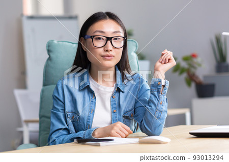 Portrait of a young Asian student girl in glasses and denim clothes, sitting at a desk with a notebook and a pen, looking at the camera, smiling. 93013294