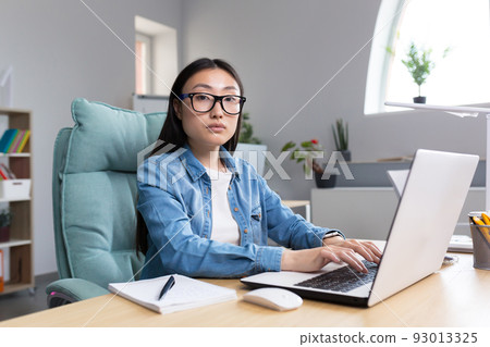 Distance Learning. A young Asian woman is a teacher, conducts online classes, lessons. He looks into the camera, explains, tells. Sitting in glasses and jeans at a table at a computer in a classroom 93013325