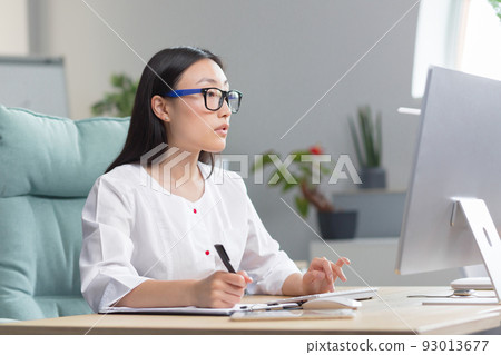 The doctor is online. A young beautiful Asian woman doctor conducts an online consultation. He is sitting in a modern office at a work table, talking on a video call with a patient at a computer. 93013677