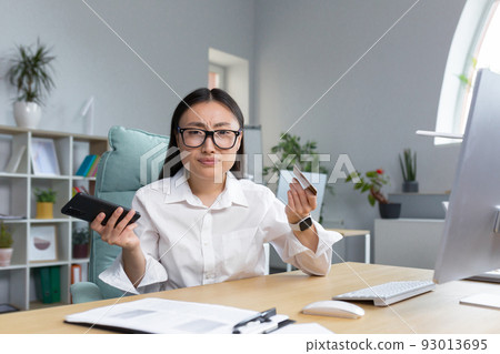 Fraud. Upset young beautiful Asian woman holding phone and credit card. A disappointed young lady lost her life savings to a scam. Sitting at the desk in the office Fraud. Upset young beautiful Asian woman holding phone and credit card. A disappointed young lady lost her life savings to a scam. Sitting at the desk in the office 93013695