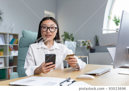 Online shopping. A young beautiful Asian woman holds a mobile phone and a credit card in her hand, makes an order on the phone, pays by card.. Sitting at a table in a modern office. Looks into the 93013696