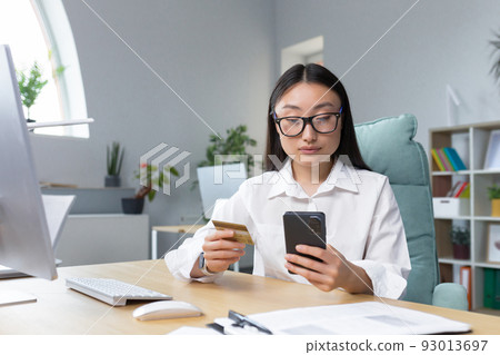 Online shopping. A young beautiful Asian woman holds a mobile phone and a credit card in her hand, makes an order on the phone, pays by card.. Sitting at a table in a modern office. Online shopping. A young beautiful Asian woman holds a mobile phone and a credit card in her hand, makes an order on the phone, pays by card.. Sitting at a table in a modern office. 93013697
