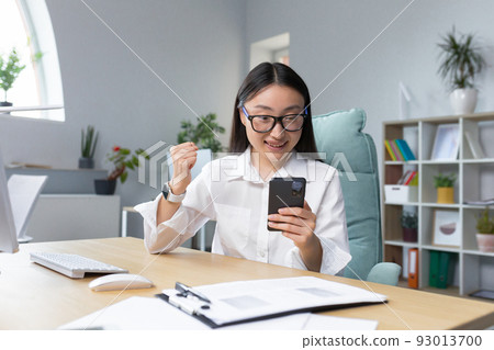 Excited beautiful young asian business woman in glasses win after achievement reading smart phone while sitting on desktop in office. She is happy, rejoices, shows a winning gesture with her hand Excited beautiful young asian business woman in glasses win after achievement reading smart phone while sitting on desktop in office. She is happy, rejoices, shows a winning gesture with her hand 93013700