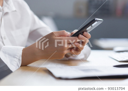 Close-up photo. The hands of a young woman in a white shirt are holding a mobile phone, typing a message at the table. Close-up photo. The hands of a young woman in a white shirt are holding a mobile phone, typing a message at the table. 93013704