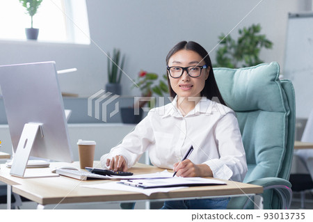 Workaholic. A young Asian business woman, sitting at the table drinking coffee to go, a paper cup of coffee is standing on the desktop near the computer. He looks at the camera, smiles. 93013735