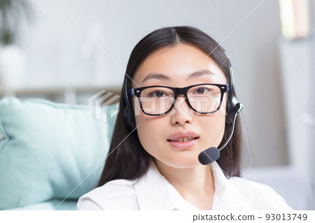 Portrait of a young Asian woman in headphones with a microphone. Works as a consultant in a call center, answers calls. Sitting at the table in glasses at the computer,looking at the camera, smiling. Portrait of a young Asian woman in headphones with a microphone. Works as a consultant in a call center, answers calls. Sitting at the table in glasses at the computer,looking at the camera, smiling. 93013749