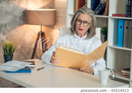 Gray-haired woman sitting at the table and working with project documentation Gray-haired woman sitting at the table and working with project documentation 93014013
