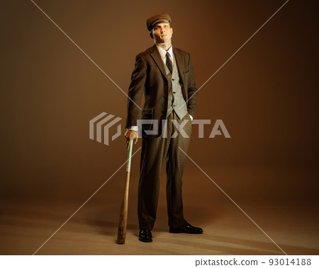 Retro style portrait of young man in image of english gangster, businessman wearing suit and cap standing isolated over dark vintage background. Retro style portrait of young man in image of english gangster, businessman wearing suit and cap standing isolated over dark vintage background. 93014188