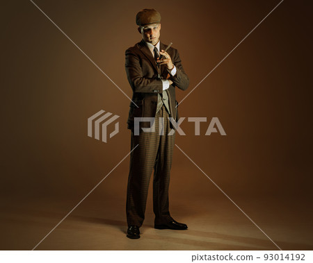 Retro style portrait of young man in image of english gangster, businessman wearing suit and cap standing isolated over dark vintage background. 93014192