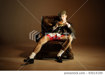 Young man, retro 1920s style english gangster jacket, shorts and cap tasting fast food isolated over dark vintage background. Business, family, art, fashion Young man, retro 1920s style english gangster jacket, shorts and cap tasting fast food isolated over dark vintage background. Business, family, art, fashion 93014199