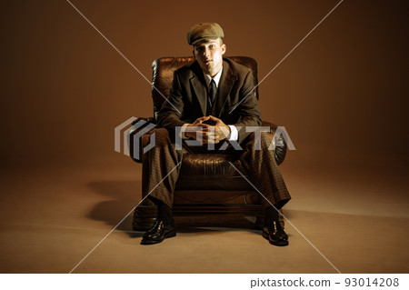 Cinematic portrait of young man, retro 1920s style english gangster wearing suit and cap isolated over dark vintage background. Business, family, art, fashion 93014208