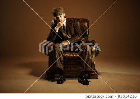 Cinematic portrait of young man, retro 1920s style english gangster wearing suit and cap isolated over dark vintage background. Business, family, art, fashion 93014210