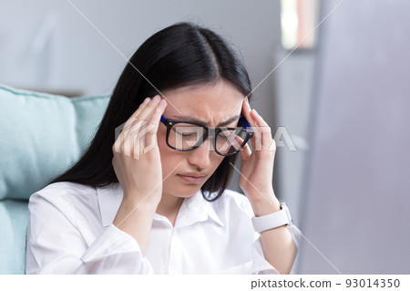 Exhaustion at work. A young Asian woman in glasses holds her head, feels pain, grimaces. Sitting in the office at the desk, working at the computer, exhausted. Businesswoman, accountant, freelancer. Exhaustion at work. A young Asian woman in glasses holds her head, feels pain, grimaces. Sitting in the office at the desk, working at the computer, exhausted. Businesswoman, accountant, freelancer. 93014350