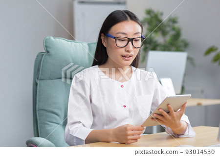 Young beautiful Asian female doctor working in modern clinic, using tablet for video calls and online consultation of patients. Young beautiful Asian female doctor working in modern clinic, using tablet for video calls and online consultation of patients. 93014530